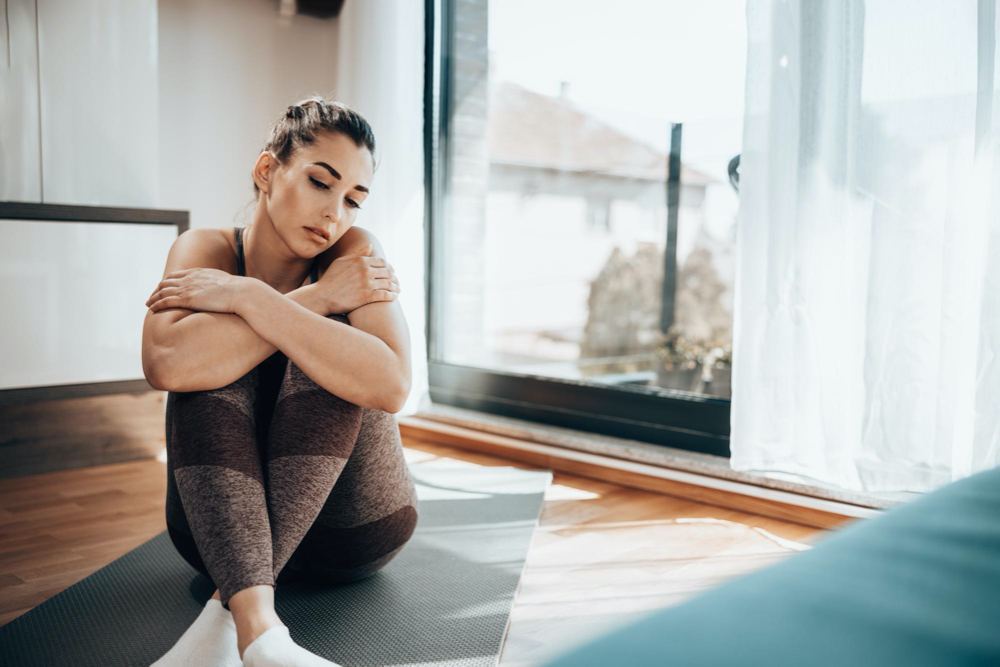 Pensive young woman is sitting on the yoga mat and preparing for training at home.