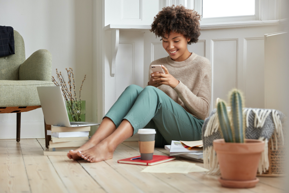 positive attractive black lady holding smart phone while drinking coffee at home.