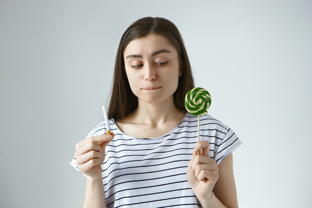 young brunette woman in striped top holding lollipop in one hand and cigarette in other,