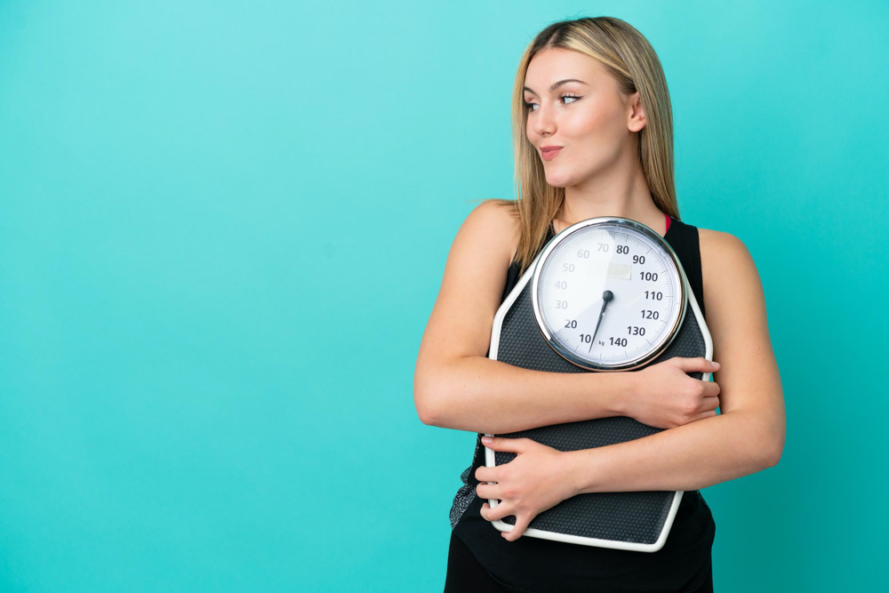 young caucasian woman isolated on blue background with weighing machine and looking side